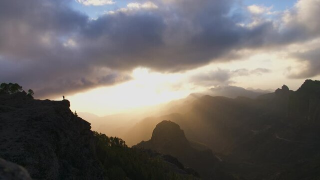 Dramatic timelapse of clouds moving over the mountains of Gran Canaria. Golden sunrays beam through the cloudy sky at sunset, illuminating the landscape