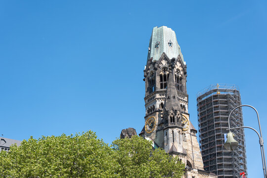Kaiser Wilhelm Memorial Church rises above London plane trees in Berlin. Scaffolding and clear blue sky frame the historic tower.