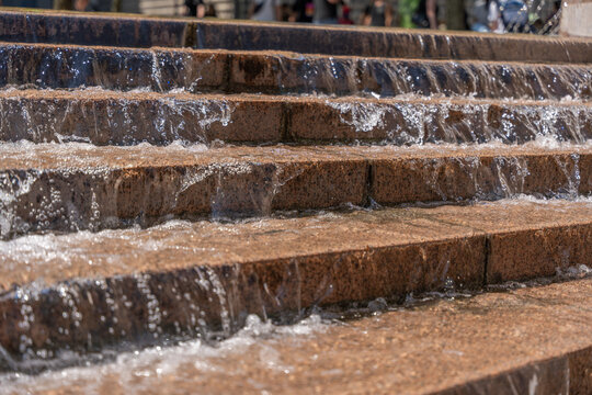 Clear water flows across layered stone fountain tiers under bright sunlight. The close composition emphasizes sparkling spray, wet texture and repeating horizontal lines.