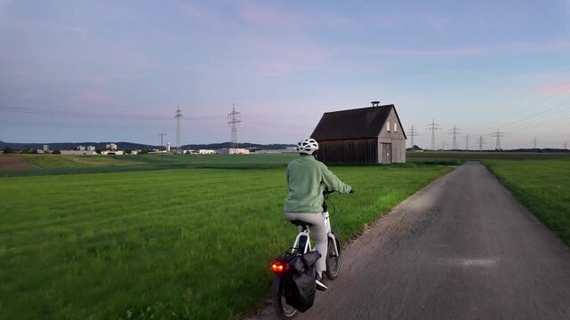 Woman cyclist riding electric bicycle on rural road in evening countryside  