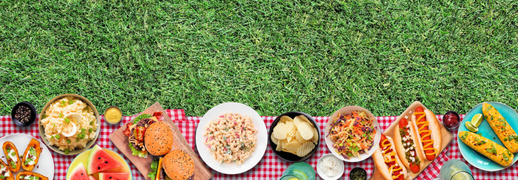 Outdoor summer food BBQ bottom border with hot dogs, hamburger and sides. Above view on a red and white check cloth and grass banner background.