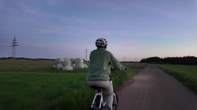 Woman cyclist riding electric bicycle on rural road in evening countryside  
