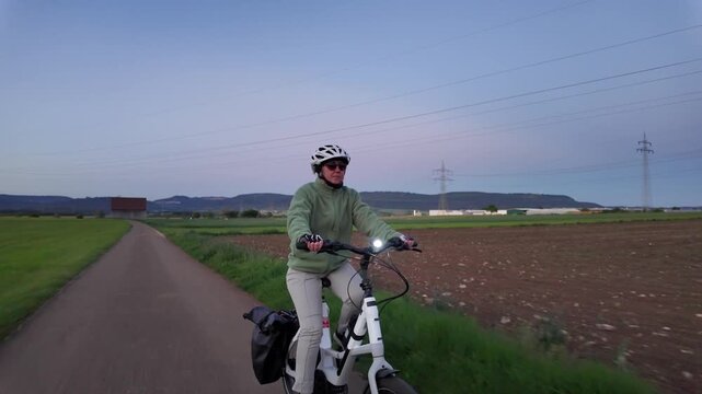 Woman cyclist riding electric bicycle on rural road in evening countryside  