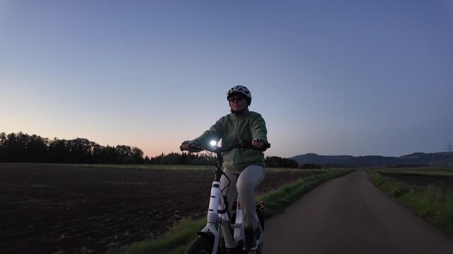 Woman cyclist riding electric bicycle on rural road in evening countryside  