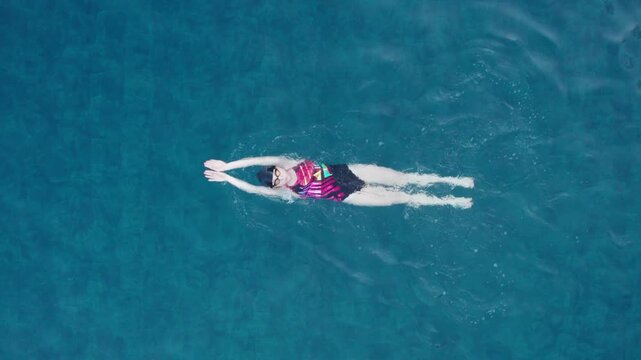 Top view of woman swimming breaststroke on back in outdoor pool. Stock video shows symmetrical leg kick, streamlined glide, and controlled backstroke style fitness movement