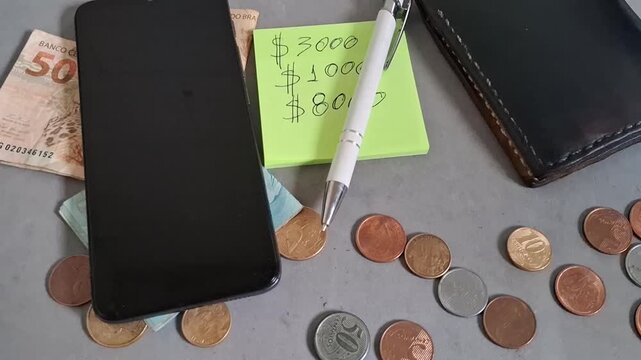 Smartphone and wallet with scattered brazilian coins and a note detailing various financial savings goals