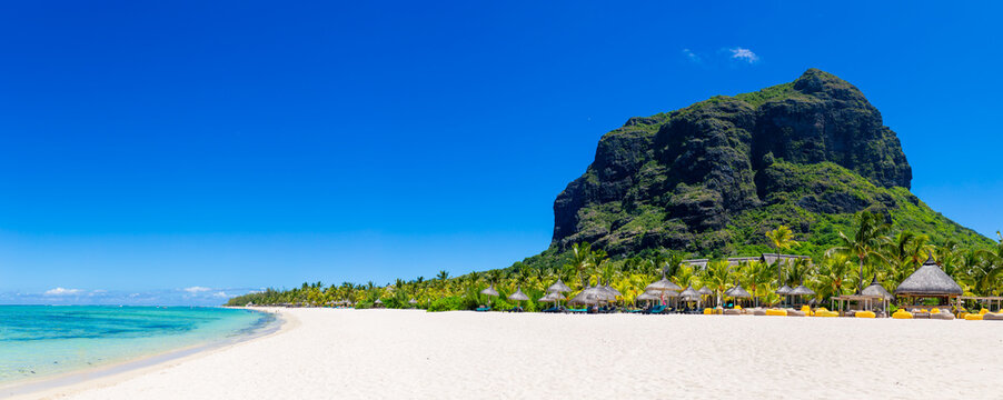 A luxurious white sand beach with palm trees, sun loungers, and umbrellas in Mauritius. A beautiful tropical beach with Le Morne Mountain as a backdrop. The coastline of an African island.