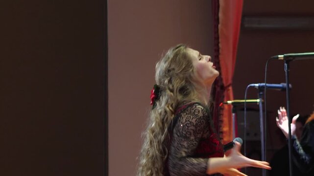 Expressive young female choral director with long curly hair wearing gorgeous red gown dynamically directing mixed vocal ensemble with emotional hand