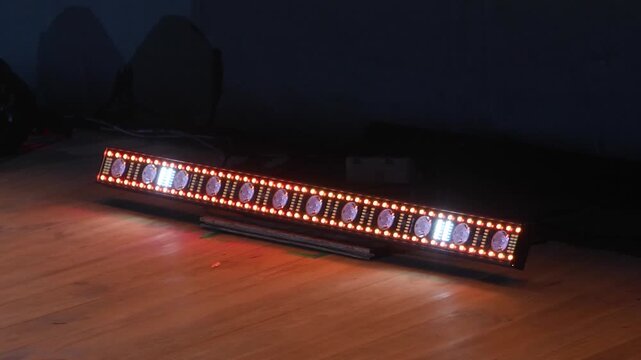 Portable linear LED fixture emitting pulsing orange and flashing white light patterns placed on dark wooden floor for professional stage