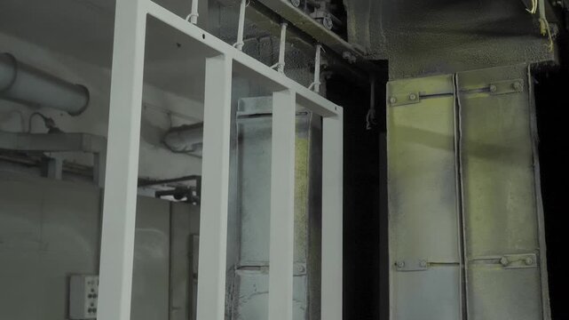 White painted rectangular metal frame component hangs from hooks on automated overhead conveyor belt mechanism moving slowly through dark industrial workshop area.