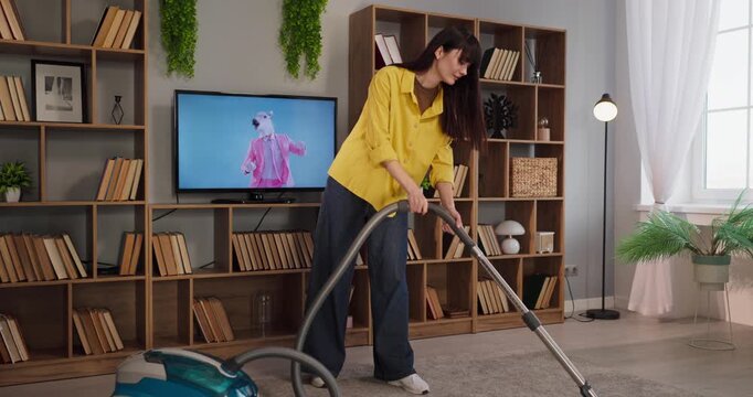 Woman vacuuming carpet in home livingroom. She does housework with an appliance, keeping hygiene and cleaning while the television is on. Concept domestic cleanliness and everyday chores.