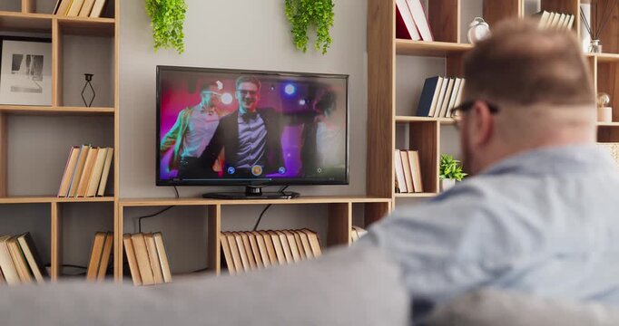 Overweight man watching television from sofa at home. Rear view shows a relaxed viewer enjoying entertainment in a living room with plants. Concept shows home leisure and deep relaxation.