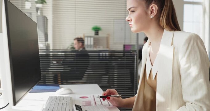 Woman analyst in office uses computer analysis. She reviews finance data and marks a report, planning next steps at a clean workspace preparing insights. Clear data driven planning concept.