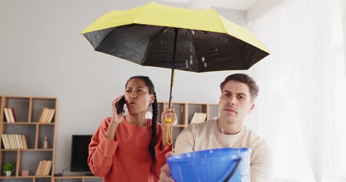 Couple manages leak with umbrella, bucket, phone. At home a man catches drips in a bucket as a woman calls a plumber about the leaky ceiling. Concept highlights home water leak emergency.