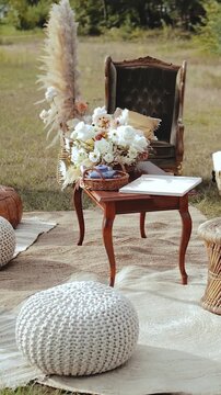 Vertical video. Cozy outdoor wedding lounge area with vintage armchair, wooden table, floral arrangement and soft knitted poufs on natural rugs. Elegant boho ceremony decor in daylight. Filmed in 4K