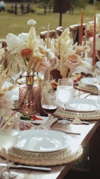 Vertical video. Elegant outdoor wedding table decorated with floral arrangements, pampas grass and stylish glass vases in warm daylight. Detailed shot of boho table setting, white flowers and luxury