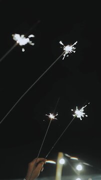 Vertical video. People holding sparkling sparklers during outdoor evening celebration under dark night sky. Close-up of glowing lights and festive atmosphere at romantic wedding party or joyful event