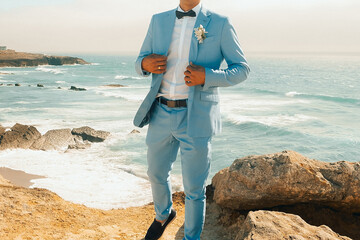 Groom in light blue suit stands on rocky beach by ocean during sunset with waves crashing in background.  man in  light blue suit adjusts his outfit while standing on rocks by ocean.  © SValeriia