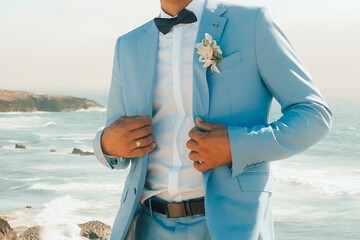 Groom in light blue suit stands on rocky beach by ocean during sunset with waves crashing in background.  man in  light blue suit adjusts his outfit while standing on rocks by ocean.  © SValeriia