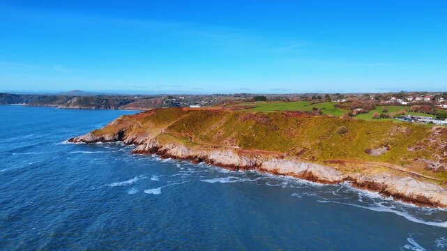 Aerial View of Rugged Welsh Coastline with Cliffs and Blue Sea. Waves break against a rocky headland topped with green and rust heathland as a coastal village sits inland under blue sky.