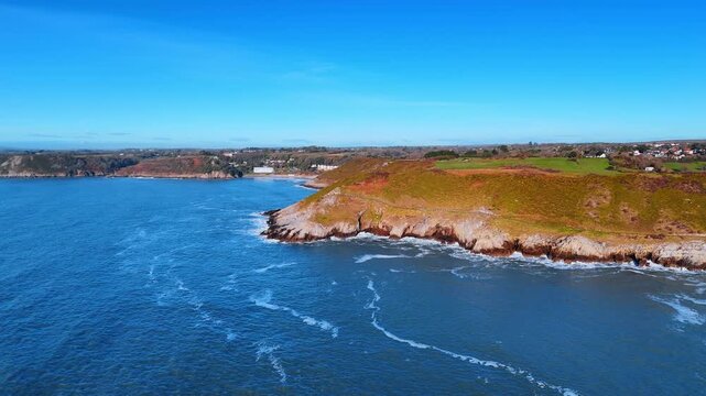 Drone View of Rocky Headland and Coastal Cliffs Along Welsh Coast. A rocky promontory covered in heathland juts into the sea as white surf breaks at its base beneath a clear blue sky.
