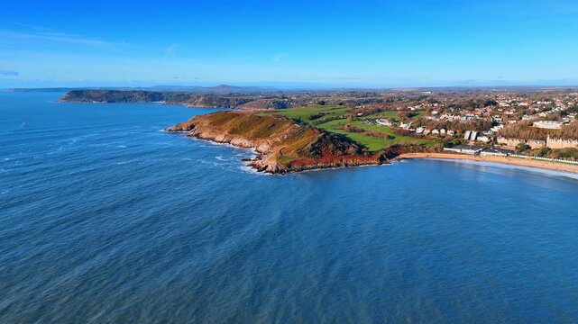 Aerial View of Coastal Headland with Town and Wide Open Sea. A rocky headland fringed by surf separates the open sea from a cliff-top town as a wide blue bay stretches to the horizon.