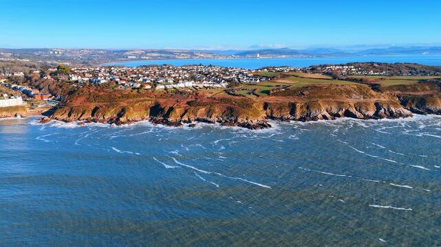 Top-Down Aerial View of Rocky Welsh Cliff Edge and Breaking Surf. A top-down view shows a rust and green cliff edge dropping to jagged dark rocks where waves break into white foam.