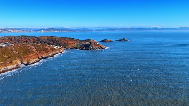 Aerial View of Welsh Headland with Beach and Coastal Village Behind. A green headland meets the sea beside a sandy cove as a coastal village and beach huts spread along the shore inland.