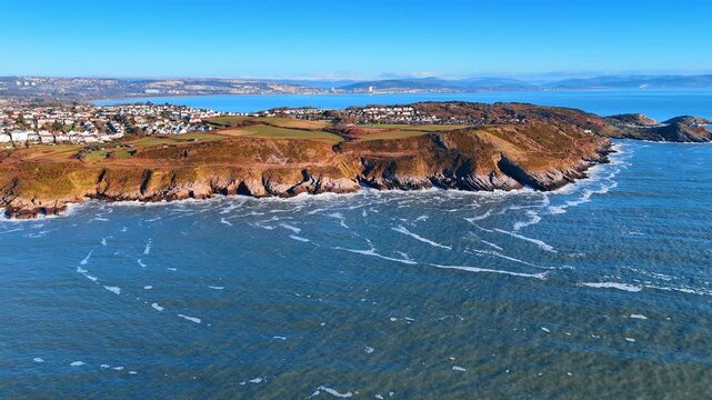 Aerial View of Promontory and Headlands Stretching Along Welsh Coast. A series of rocky headlands fringed with white surf recedes into the distance under a clear sky beside cliff-top homes.