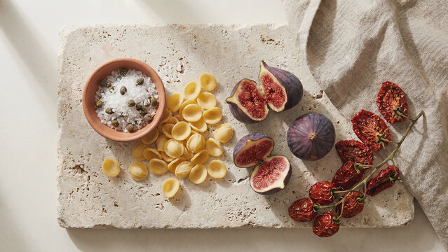 Top view of Mediterranean food ingredients on stone: orecchiette pasta, fresh figs, sun-dried tomatoes, and salt bowl with capers. Rustic culinary still life on travertine background.