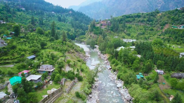 Aerial drone flying forward landscape view of the Parvati River winding through lush green Kasol valley surrounded by Himalayan mountains and riverside homes.