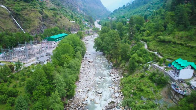 Aerial drone flying forward landscape view of the Parvati River winding through lush green Kasol valley surrounded by Himalayan mountains and transformer grid showing the electrification and developme