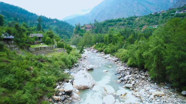 Aerial drone flying forward landscape view of the Parvati River winding through lush green Kasol valley surrounded by Himalayan mountains and riverside homes.