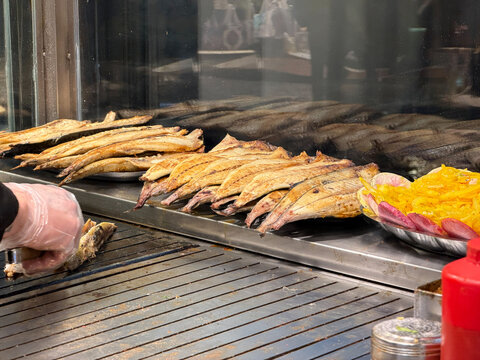 Grilled fish fillets cook on hot metal griddle for balik ekmek preparation in Istanbul street food kitchen with glass counter. Turkish food, seafood menu, culinary tourism, restaurant