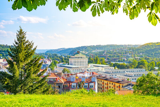 The Podol district in Prague shows a mix of buildings and green areas. People are enjoying the view from a hill. It is a sunny day with clear skies and a vibrant atmosphere.