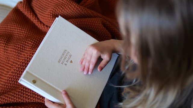 Over the shoulder view of a little girl holding a family photo album, gently tracing the embossed golden text with her fingers while resting on a cozy knitted terracotta blanket