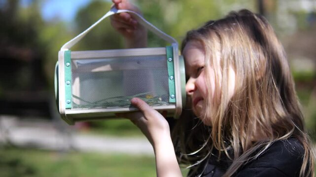 Young girl with long blonde hair carefully examining a captured bug inside a special viewing box, showing her curiosity and interest in entomology and the natural world during a sunny day outdoors