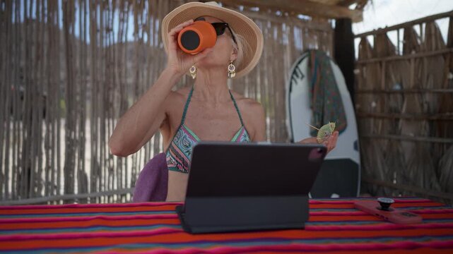 Mature woman works on computer tablet while sipping drink at beach during vacation in tropical setting under the sun.