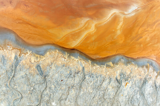 Aerial view of the abstract textures and vibrant orange acidic water of the Rio Tinto mines meeting grey mineral deposits in Minas de Riotinto, Andalusia, Spain.
