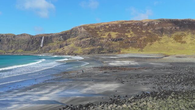 People walking on talisker bay beach with waterfall and cliffs in scotland