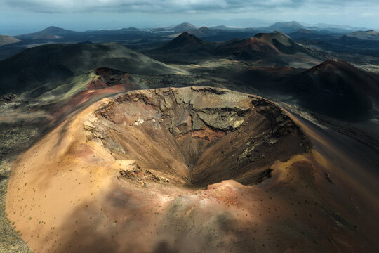Aerial view of a massive volcanic crater with reddish soil and rugged slopes in a vast lava field under a cloudy sky Lanzarote, Canarias, Spain.