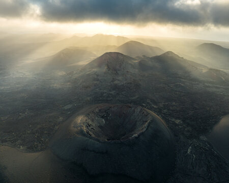 Aerial view of volcanic craters and rugged lava fields under dramatic sunbeams breaking through dark clouds in Lanzarote, Canarias, Spain.
