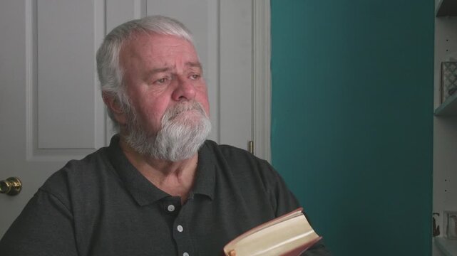 An older man sits in a quiet room looking at a book. He pauses often to think deeply about what he reads. His face shows different emotions as he reflects on the words.
