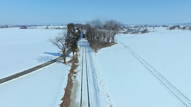 The train moves along the snowy track surrounded by a flat landscape. Trees line the way under a clear sky. It showcases the quiet charm of winter in a rural area.