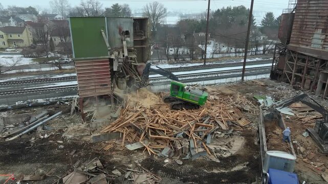 Workers are using heavy machinery to bring down a structure in a construction site. Debris and wood are scattered on the ground. The area looks cold and gray as snow is present.