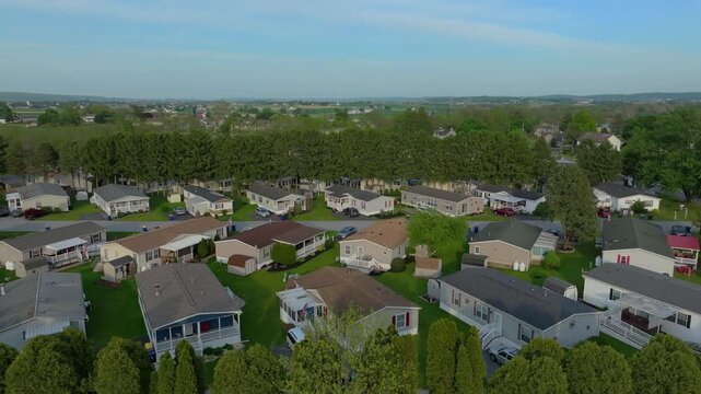 The camera shows a bird's eye view of a neighborhood. Mobile and manufactured homes are lined up with yards and trees. The sky is clear with green fields in the background. The setting calm and sunny.