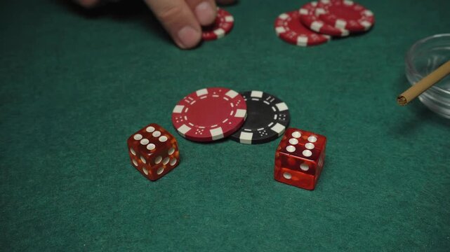 Hand placing red poker chips on a green felt table next to two orange dice during a game of chance with additional chips in the background