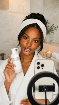 vertical UGC style skincare video of young woman in white bathrobe and headband presenting a facial cleanser pump bottle while filming on smartphone, ideal for beauty product review
