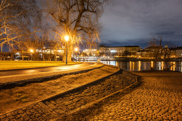 Fototapeta premium The scene shows the Vltava riverside in Prague at night. Streetlights illuminate the path along the river. Trees lean over the water. Buildings reflect on the dark surface.