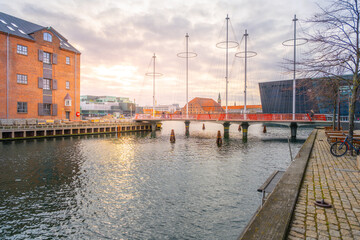 Fototapeta premium People walk and ride bicycles on Cirkelbroen. The bridge crosses Christianshavn Canal. Buildings line the water. The sun sets behind the structures creating a warm light.